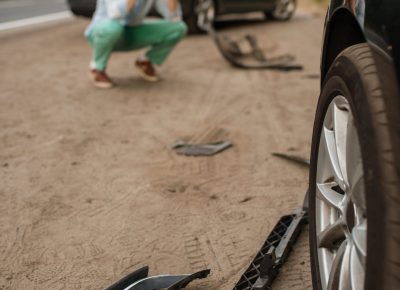 Car accident scene with broken vehicle parts scattered on the ground. A man crouches in the background near his damaged car, holding his head in distress