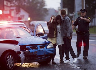Woman comforted by man at the scene of a car accident as a police officer directs traffic.